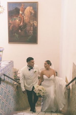 Bride and groom walk up tiled stairs holding hands beneath a equestrian painting in a warmly lit venue.