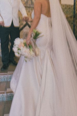 Bride in strapless white mermaid wedding gown with veil holding white floral bouquet standing beside groom in white jacket.