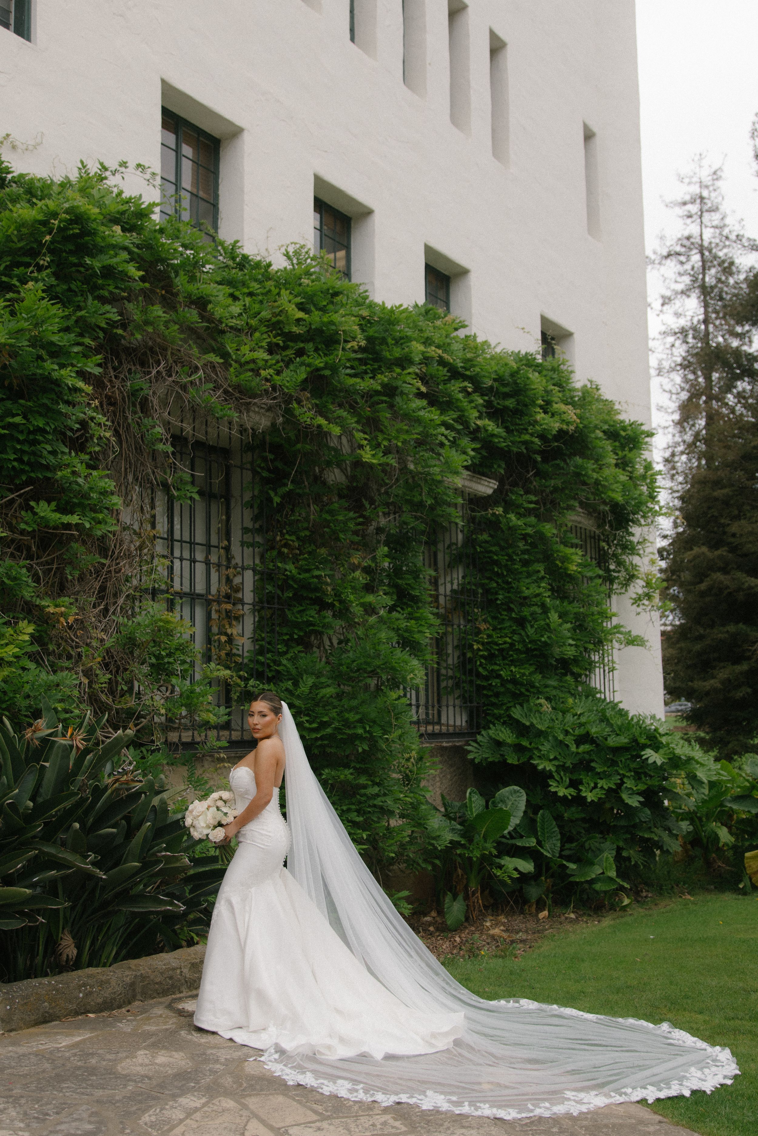 Bride in a white gown with long lace veil stands before ivy-covered white building surrounded by lush greenery.