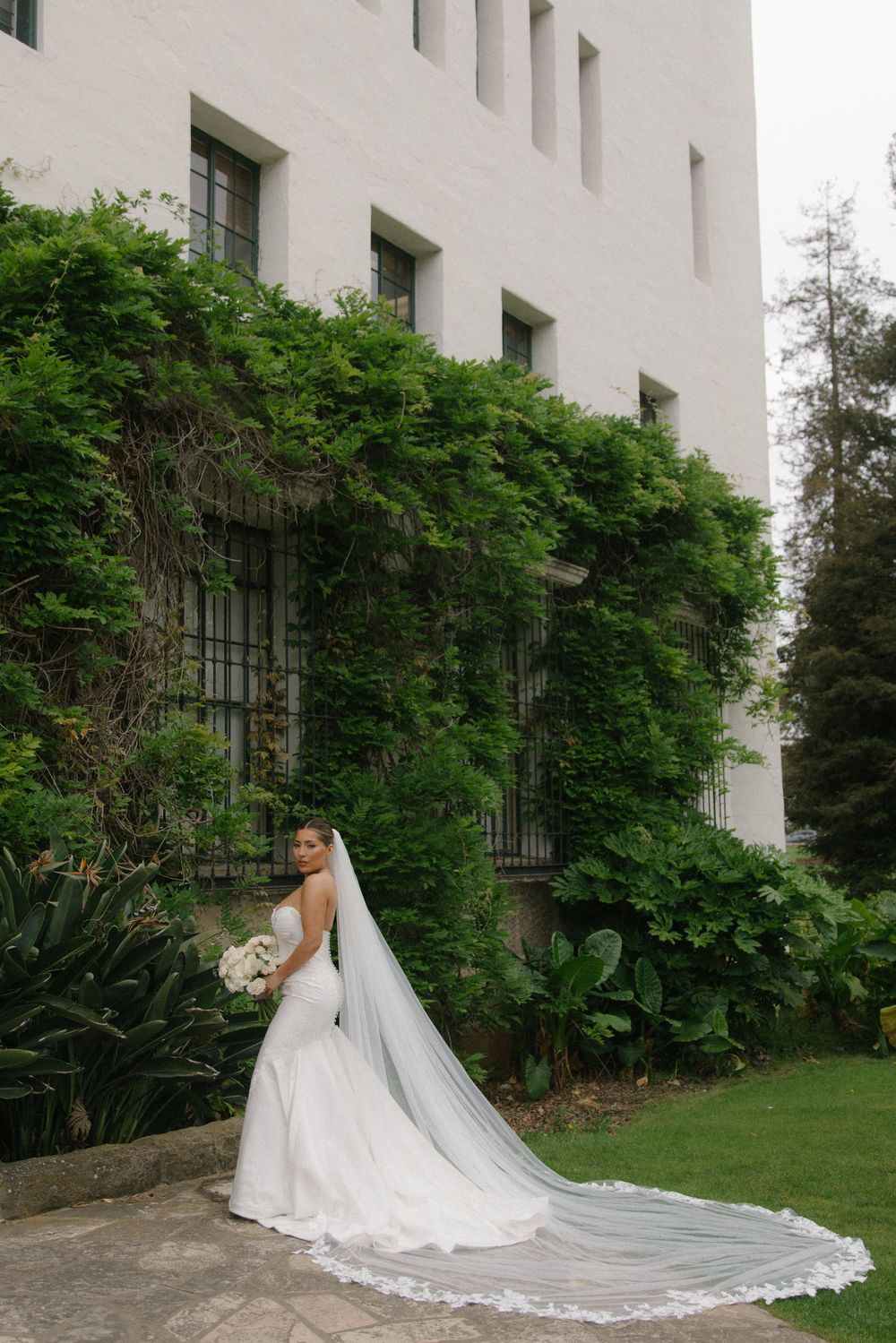 Bride in a white gown with long lace veil stands before ivy-covered white building surrounded by lush greenery.
