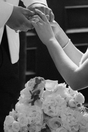 Black and white wedding photo of couple exchanging rings with a white rose bouquet in the foreground.