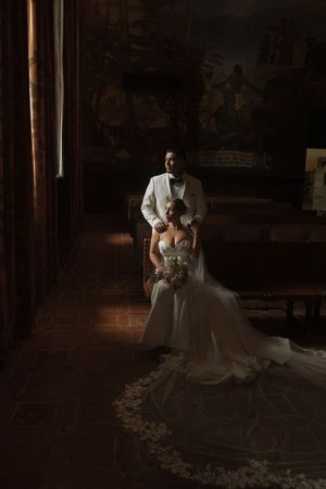 Bride in white gown seated in ornate church with groom in white suit standing behind her, dramatic lighting.