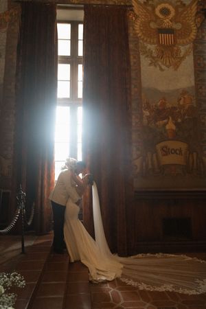 Bride and groom share a romantic kiss in an elegant hall with ornate murals and dramatic backlit window light.