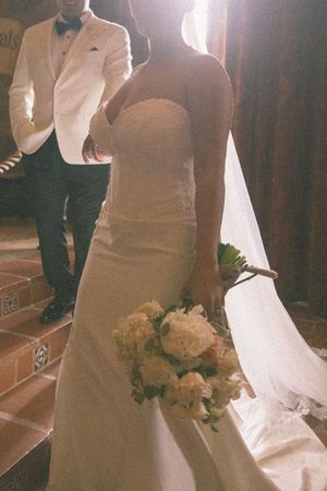 Bride in strapless white gown holds bouquet of white roses while groom in white jacket descends tiled stairs.