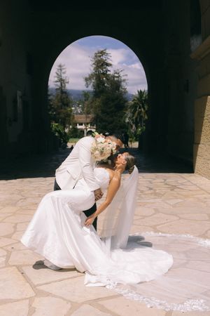Bride and groom share a romantic dip kiss under an arched doorway, bride holding white floral bouquet with veil flowing.