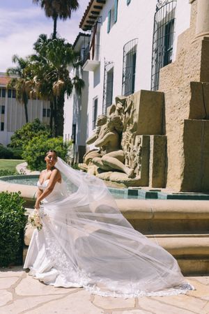 Bride in flowing white lace gown with veil billowing in wind beside ornate fountain at historic venue.