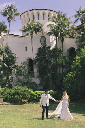 Bride and groom hold hands walking on green lawn before white Spanish-style building surrounded by tall palm trees.