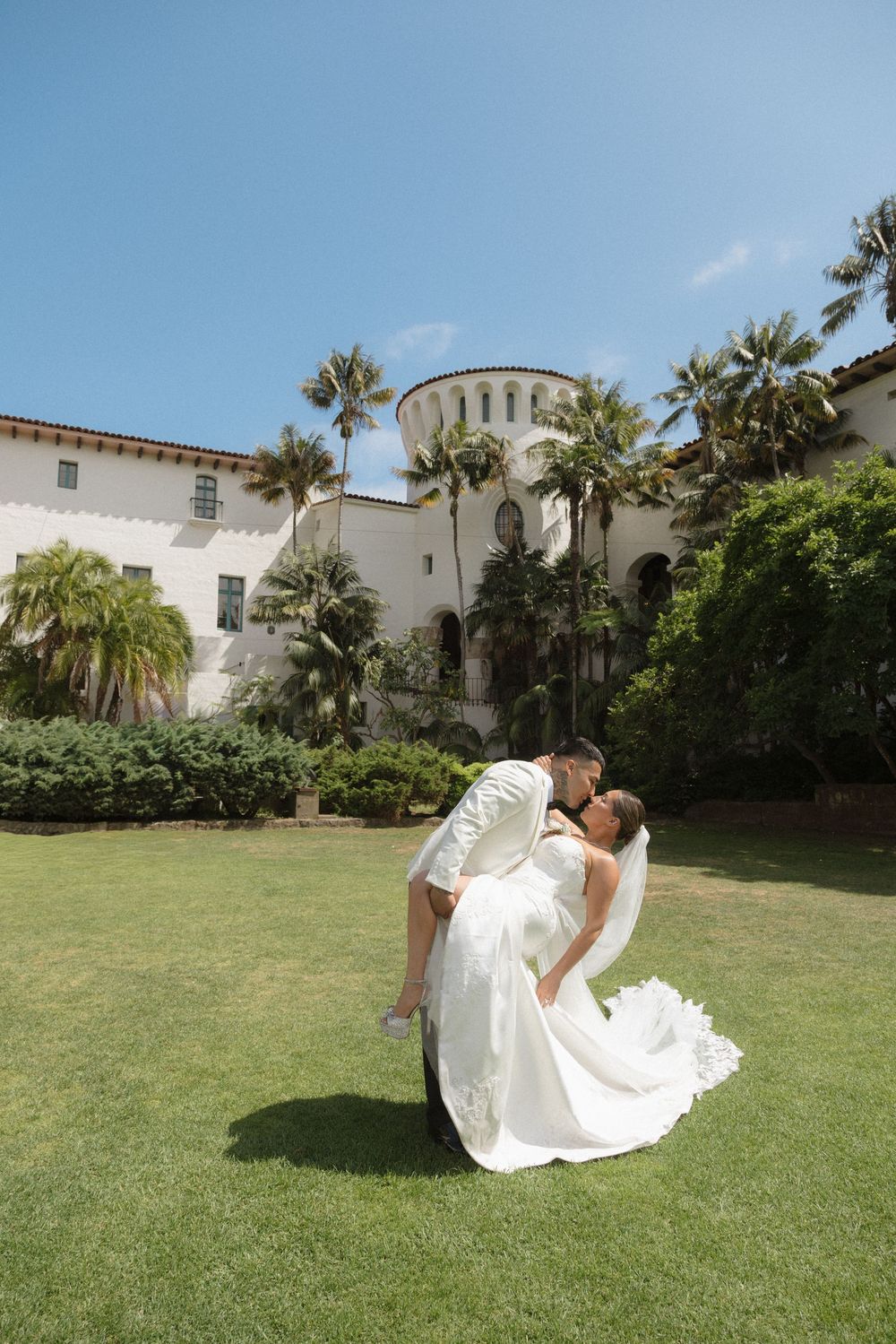 Groom dipping bride in white wedding dress on green lawn in front of white Spanish-style building.