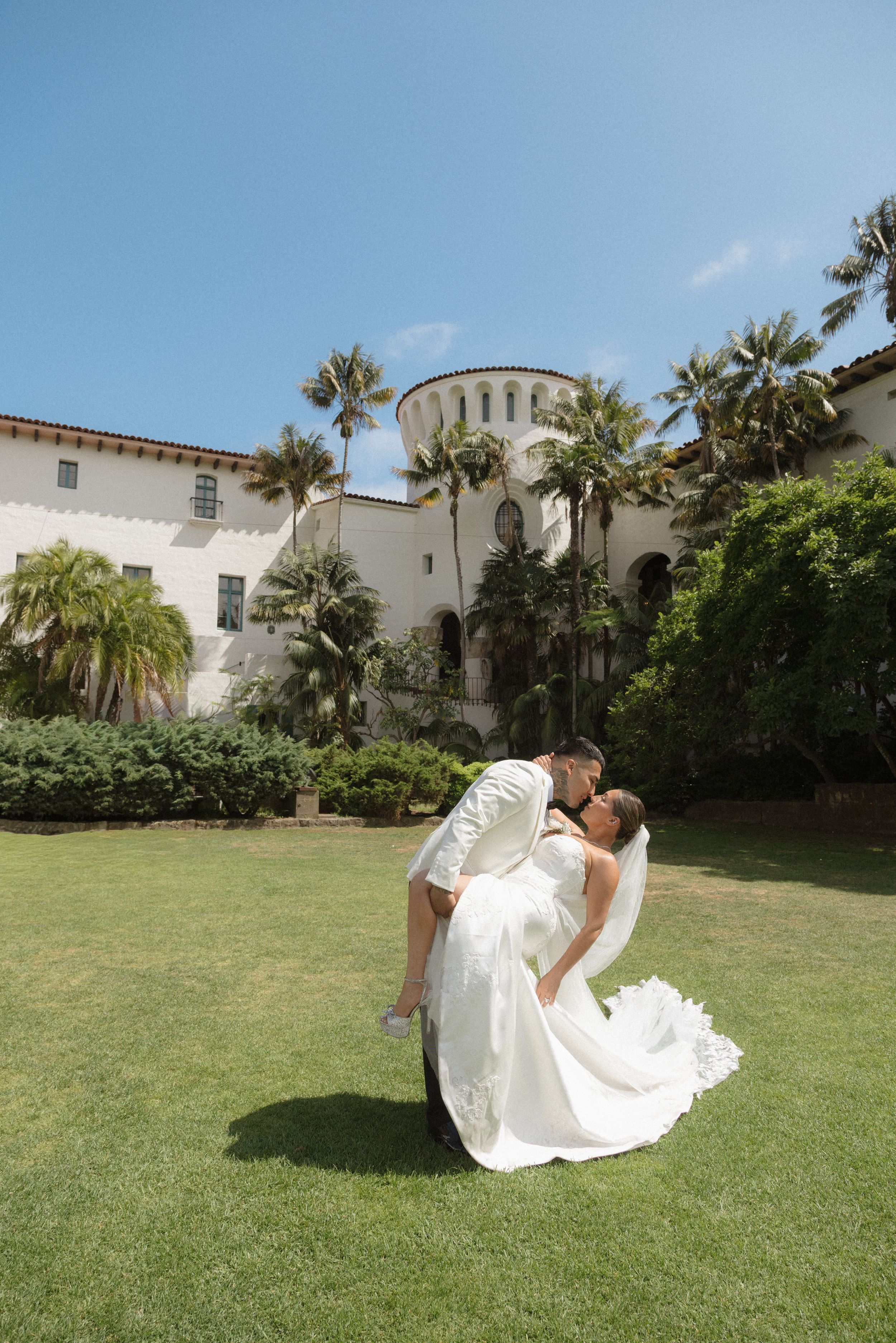 Groom dipping bride in white wedding dress on green lawn in front of white Spanish-style building.