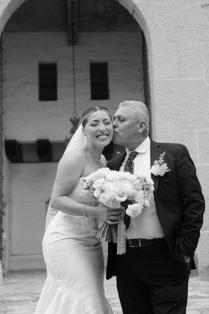 Black and white photo of a bride smiling as an older man in a suit kisses her cheek outside a stone church.