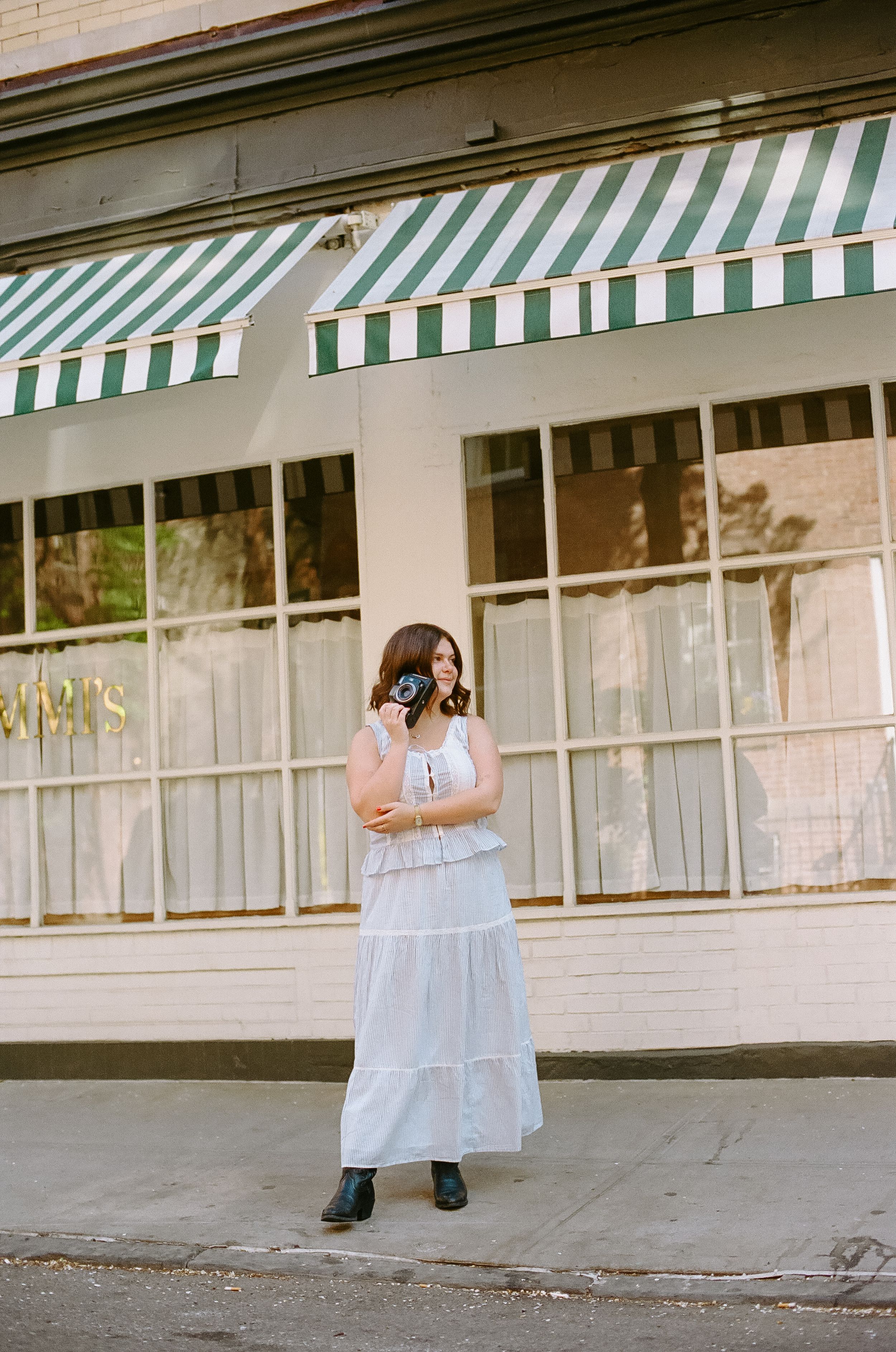 Woman in white maxi dress holding camera outside shop with green striped awnings on sunny day.