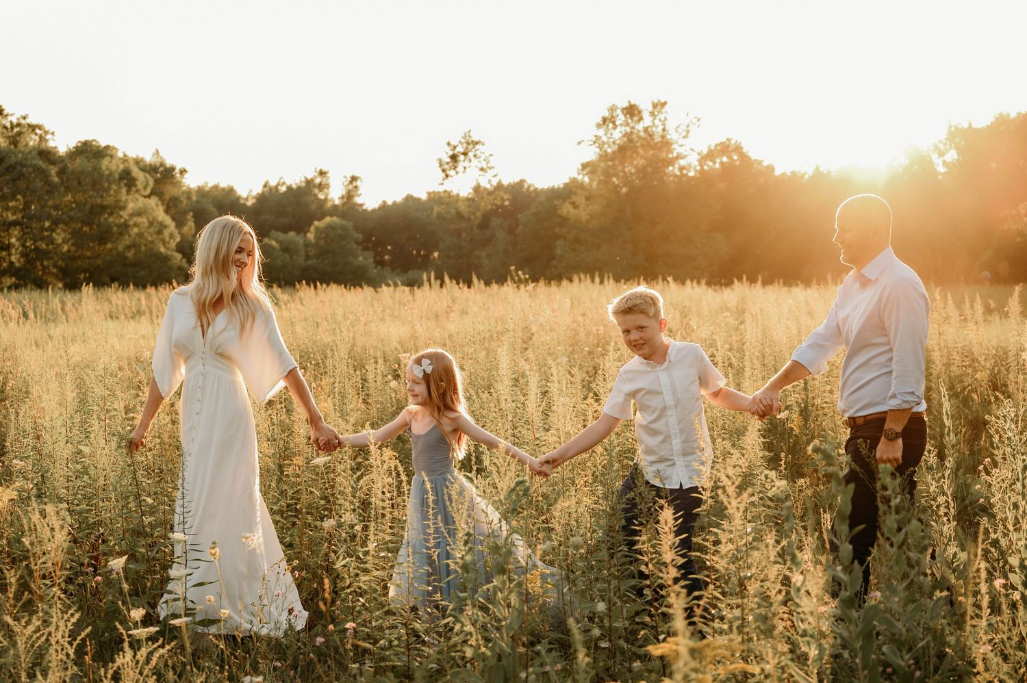 Family of four holding hands in golden sunlit meadow at sunset, parents and two children in flowing white outfits.