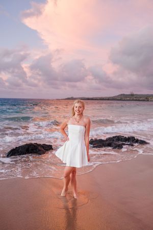 Smiling blonde woman in white strapless dress stands in ocean waves at a colorful sunset beach.