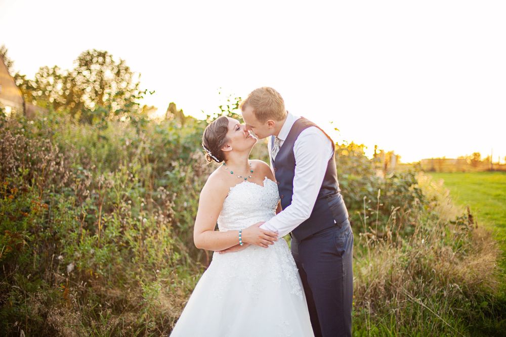 Bride and groom share a romantic kiss in a golden sunset field, bride in white gown, groom in navy suit.