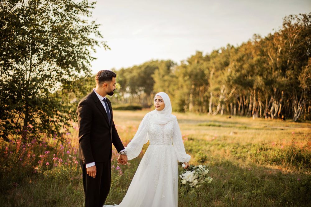 Muslim bride in white hijab and groom in black suit hold hands in a golden hour outdoor wedding photo.