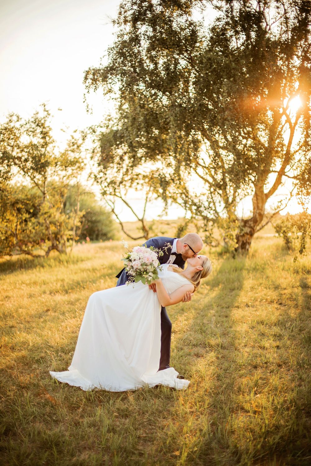 Groom dips bride for a kiss in golden sunset light among trees, bride holding pink bouquet in white gown.