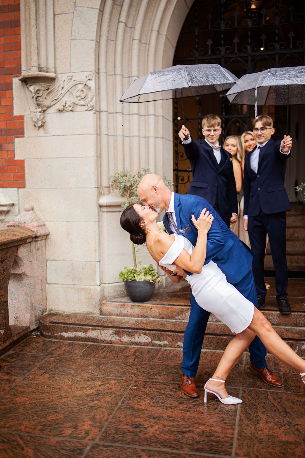 Couple shares romantic dip kiss outside stone church entrance, guests holding umbrellas in background on rainy wedding day.