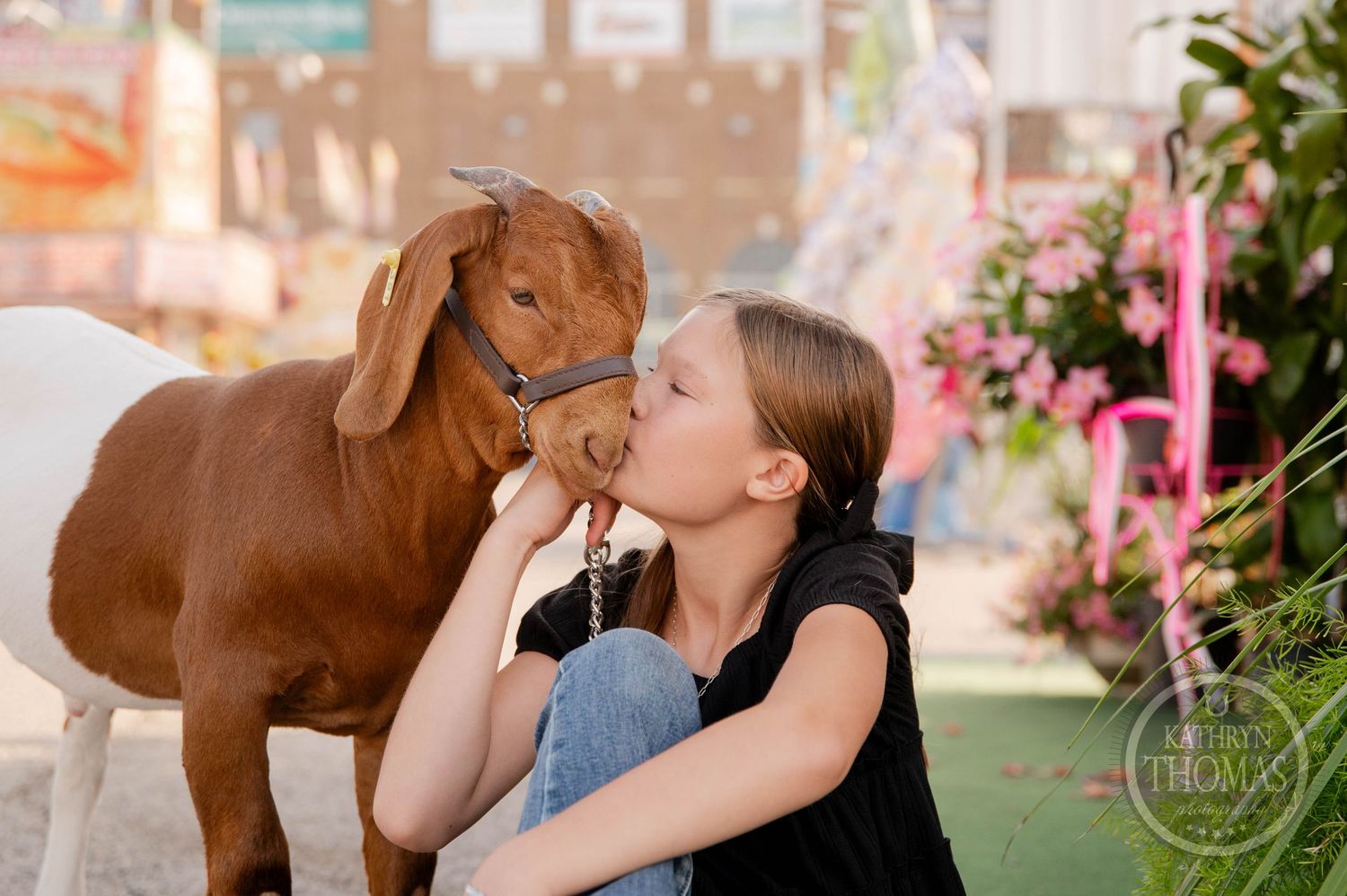A young girl kisses a brown goat on the nose at a fair, with colorful decorations in the background.