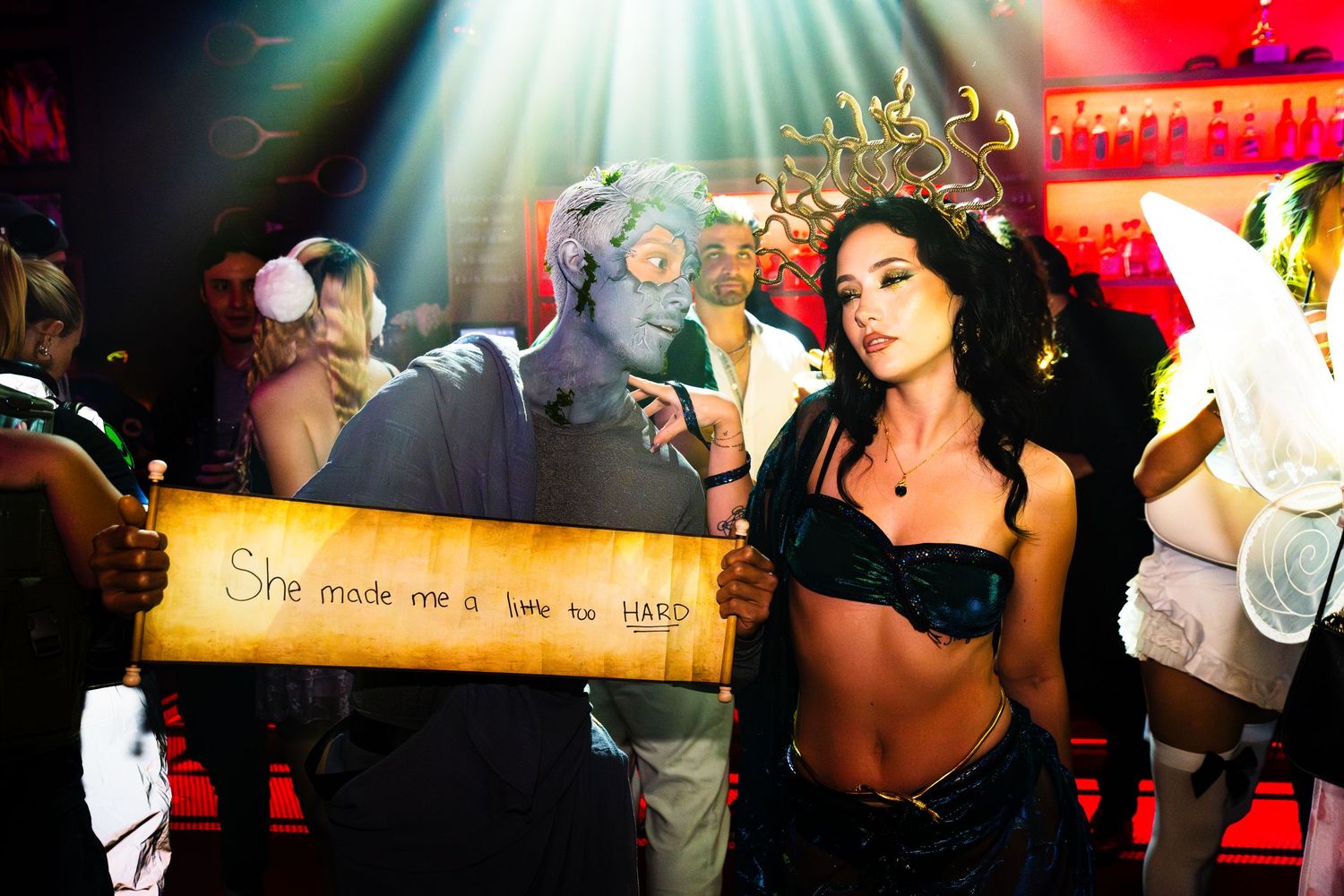 A couple in halloween costumes of medusa and a man turned into stone by her holding a sign that says "She made me a little too hard".