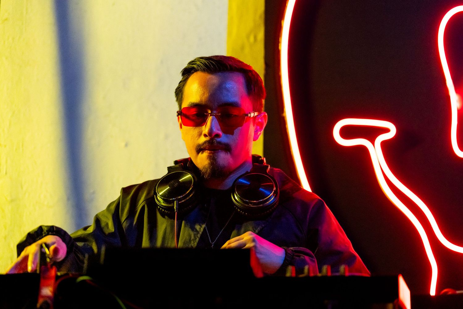 A male dj playing a set for the HDLNRS livestream in LA with a red neon glowing smiley face behind him