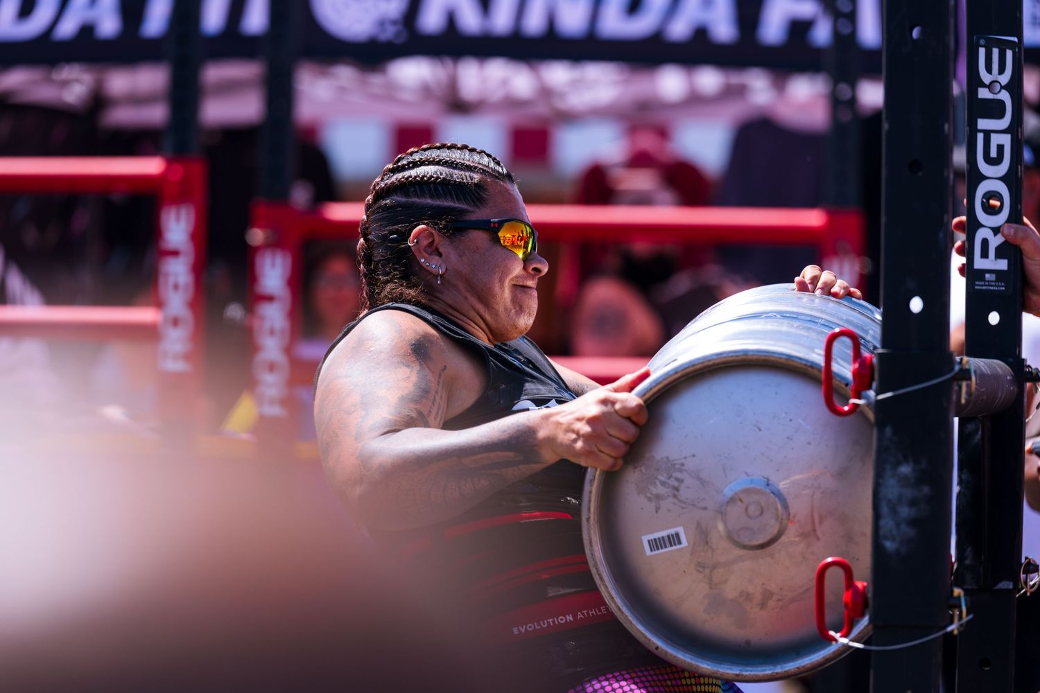A photo of a strong woman doing a keg carry at Santa Monica's Strongest Event. She attempts to lift it over the bar.