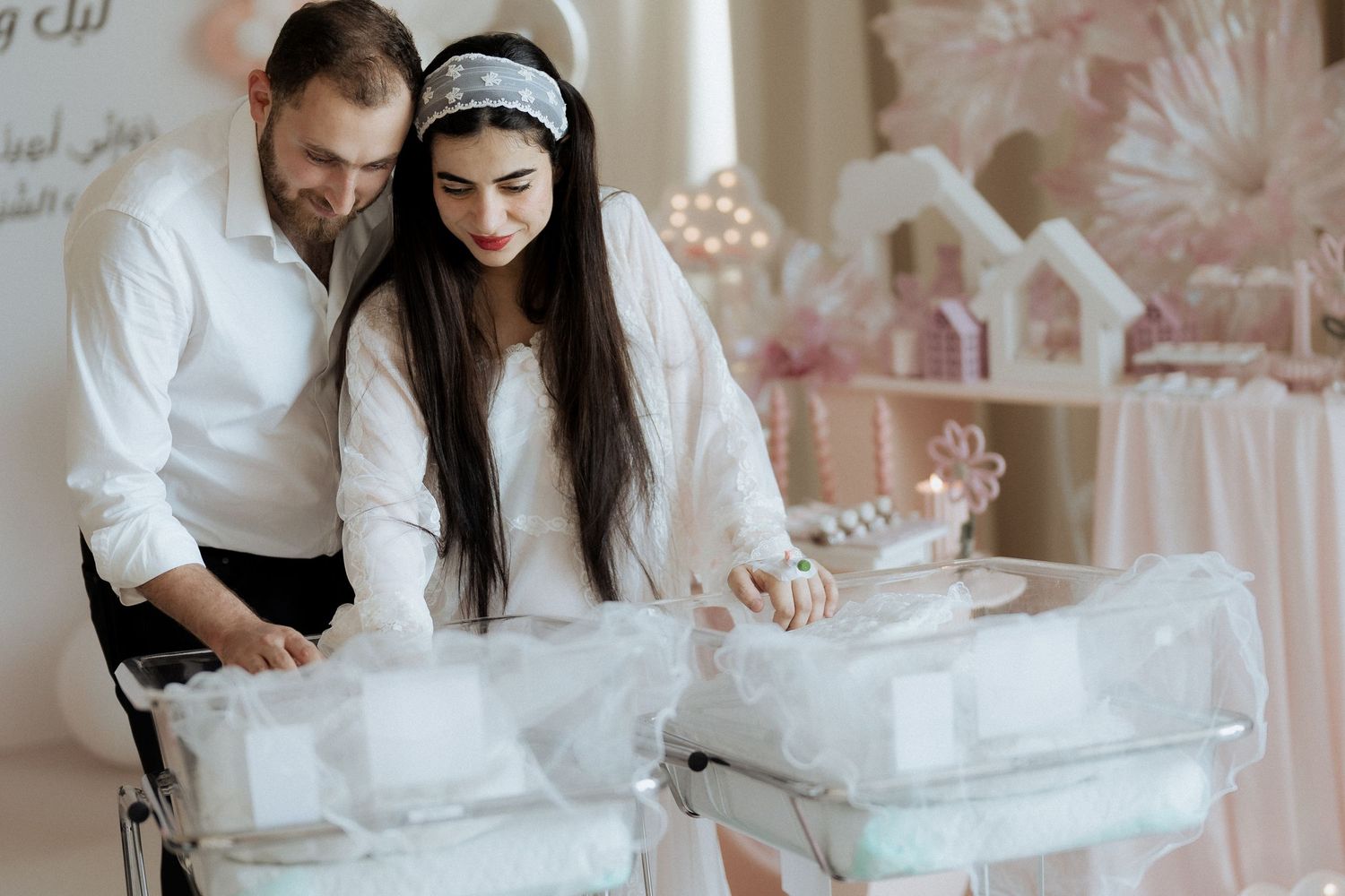 Parents in white looking down at their twin newborns in decorated bassinets at a celebration