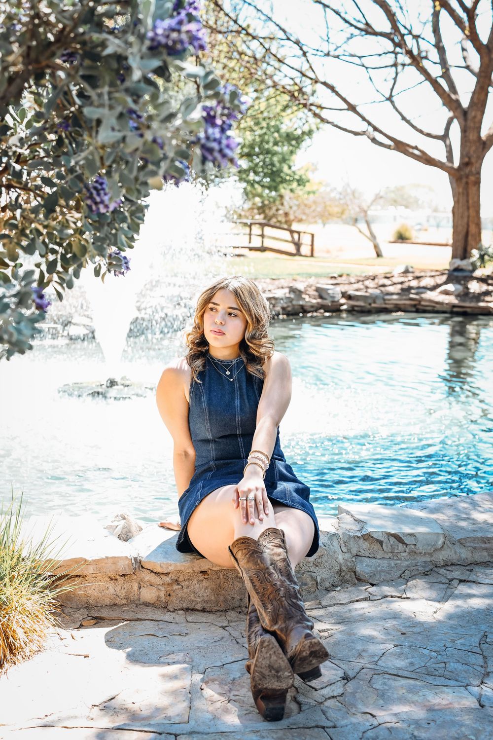 Young woman in blue dress and cowboy boots sitting by a fountain in a sunny park with purple flowering trees.