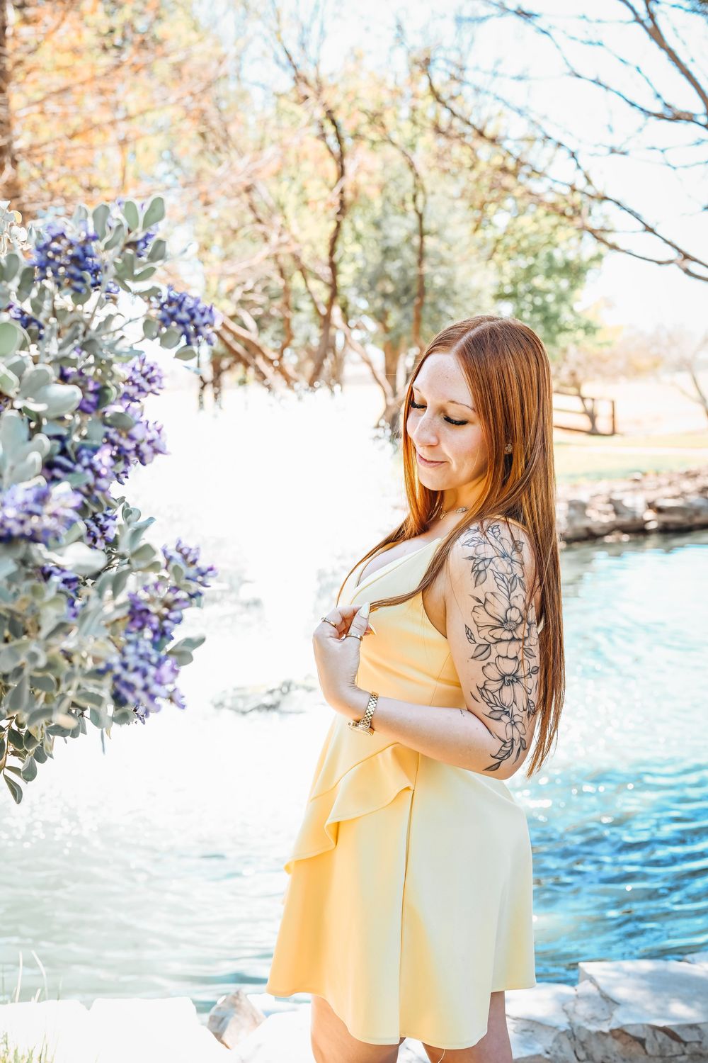 Red-haired woman with floral arm tattoo in yellow dress poses by a serene pond with purple blooming flowers.