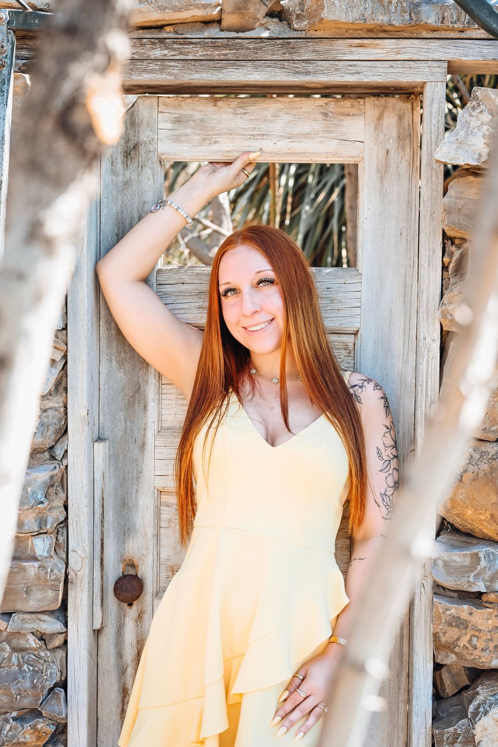 Smiling redhead woman in yellow jumpsuit posing by a rustic wooden door with stone wall backdrop.