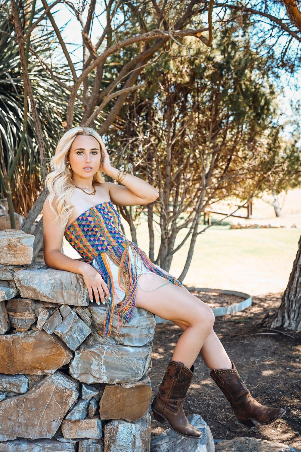 Blonde woman in colorful fringe top and brown cowboy boots sitting on stone wall outdoors.