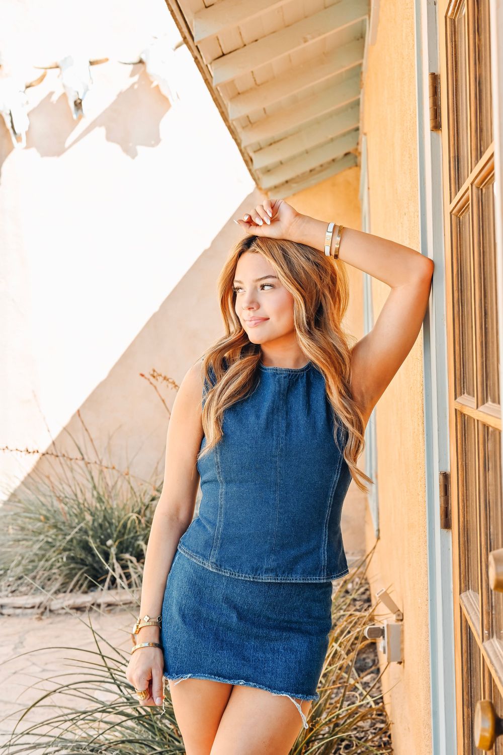 Woman in blue denim mini dress posing outdoors against a yellow wall with desert plants in background.