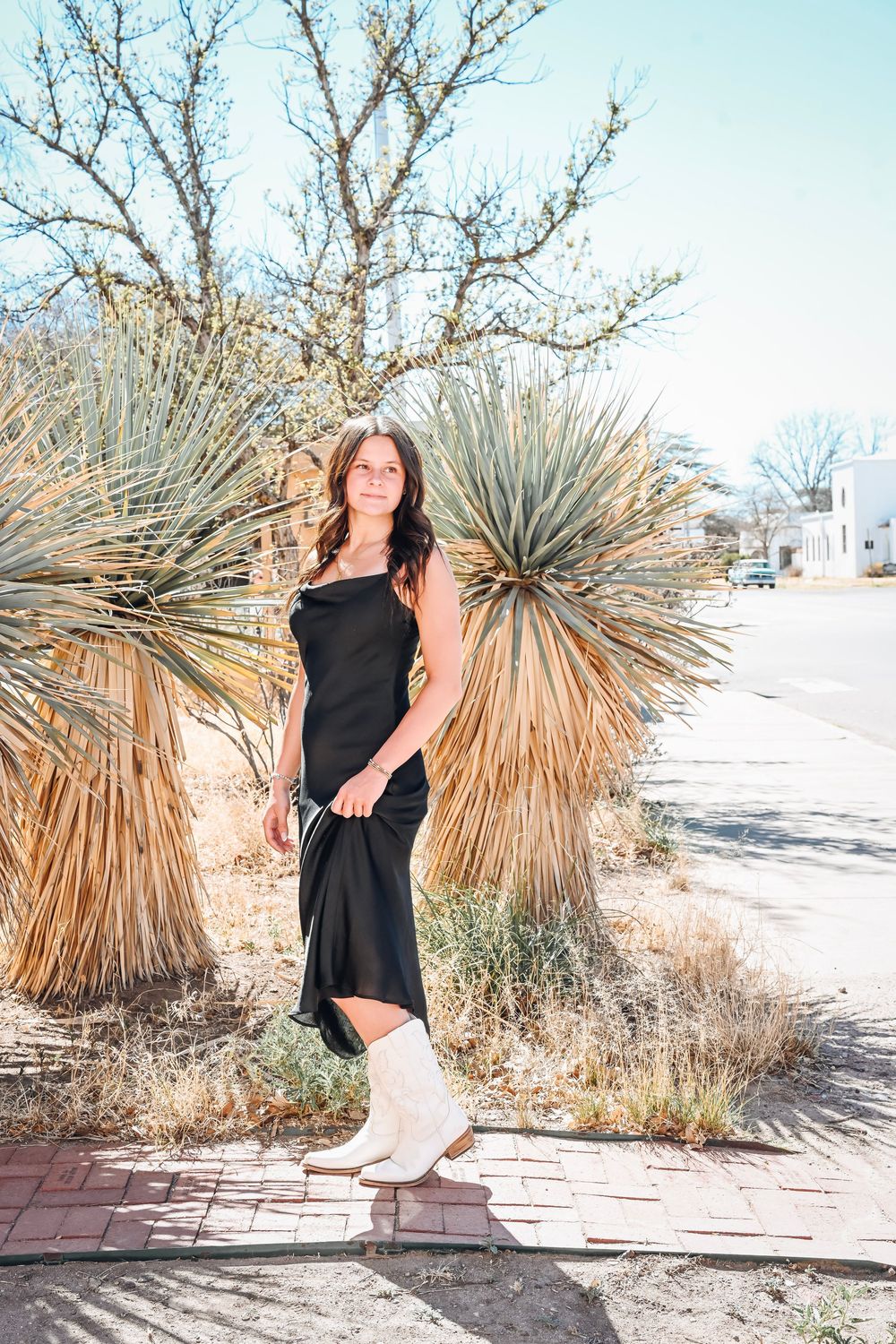 Brunette woman in black midi dress and white cowboy boots standing near desert plants outdoors.