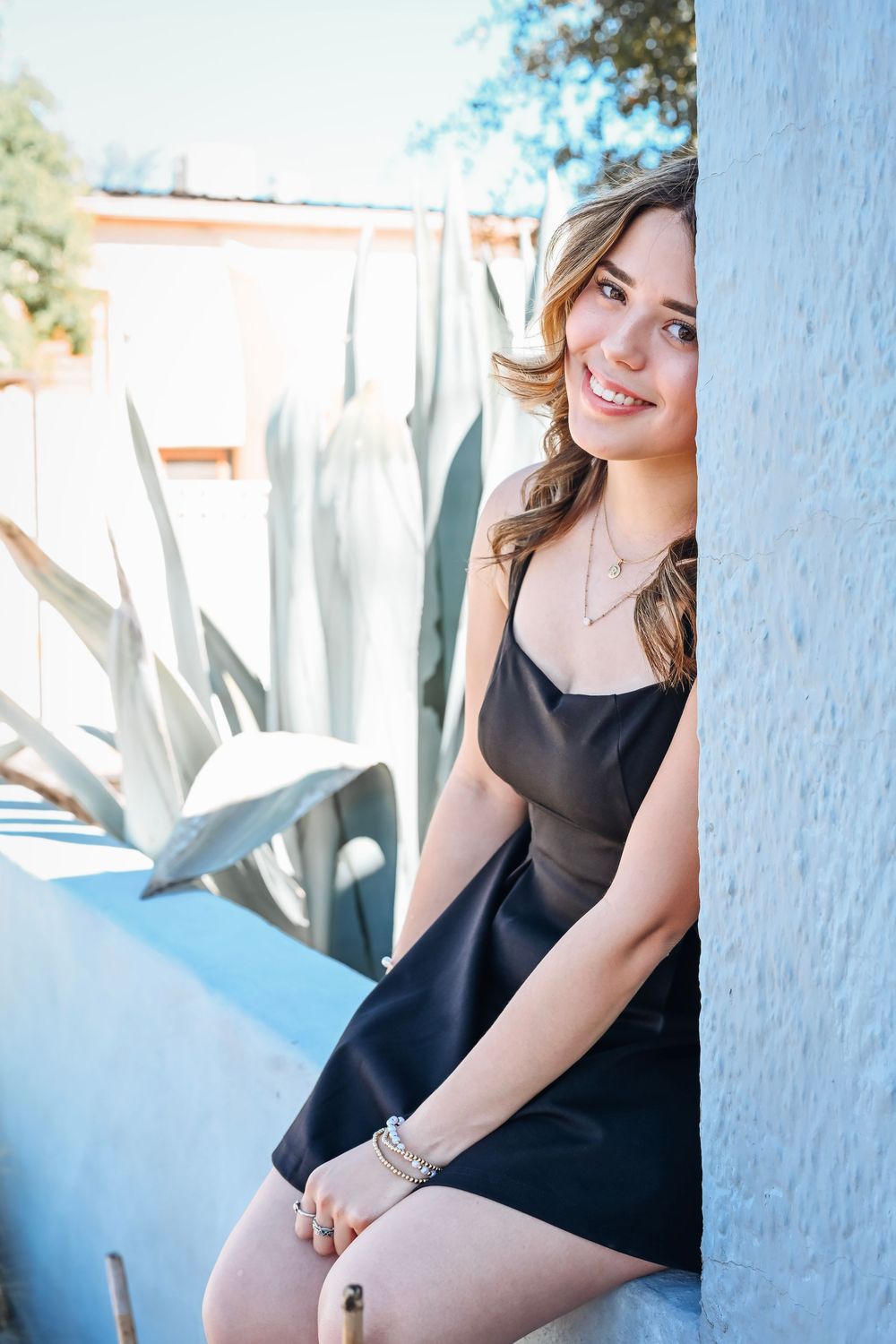 Smiling young woman in black dress sitting by blue wall with agave plants in sunny outdoor setting.