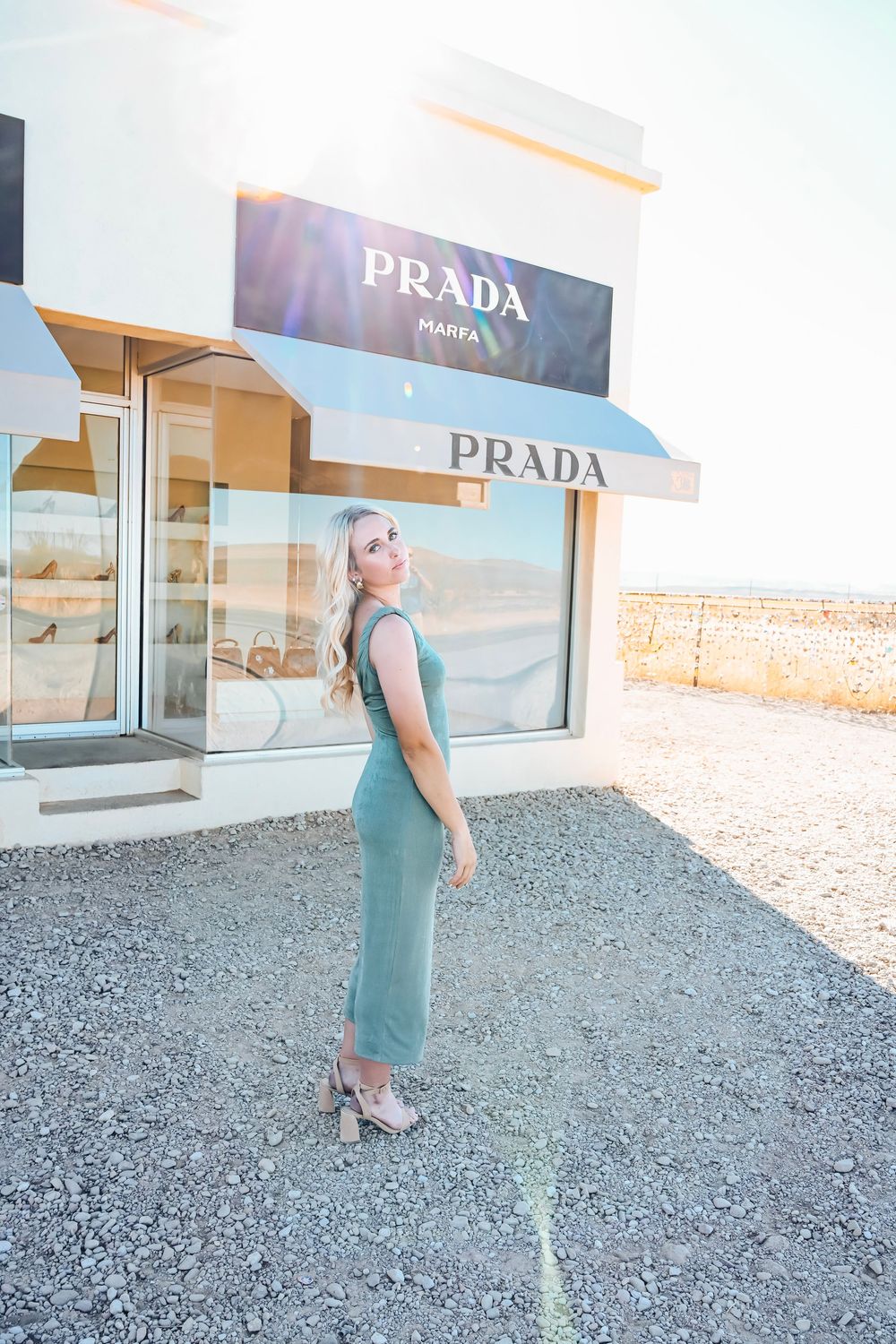 Blonde woman in teal dress posing outside Prada Marfa art installation in Texas desert.