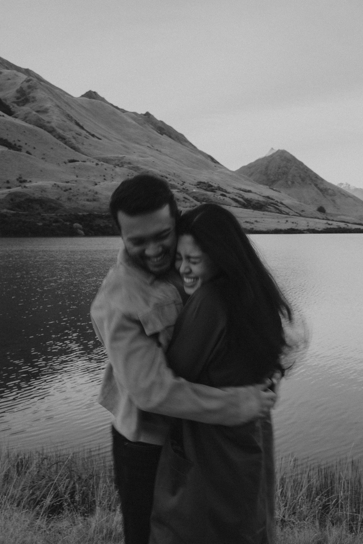 Black and white photo of a smiling couple embracing by a mountain lake, sharing a joyful moment outdoors.