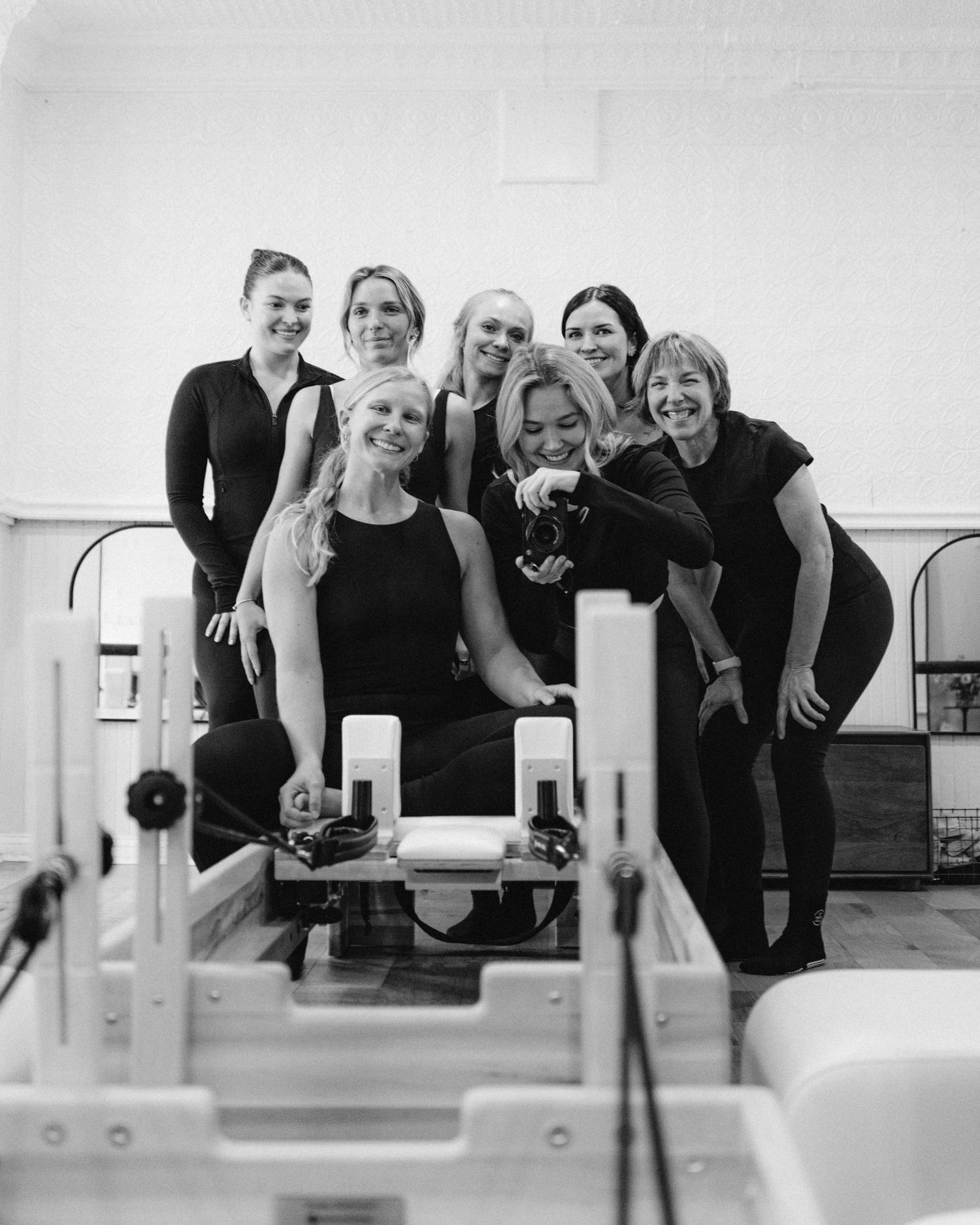 Group of seven women in black activewear posing together around a Pilates reformer machine in a fitness studio.