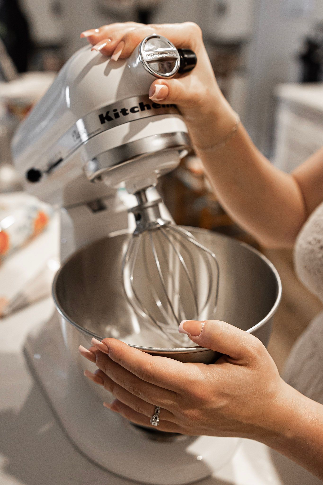 Hands adjusting a KitchenAid stand mixer with whisk attachment in a bright kitchen.