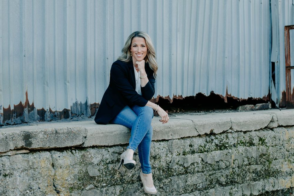 Woman in black blazer and jeans sitting on a stone wall, smiling outdoors against a rustic metal wall.