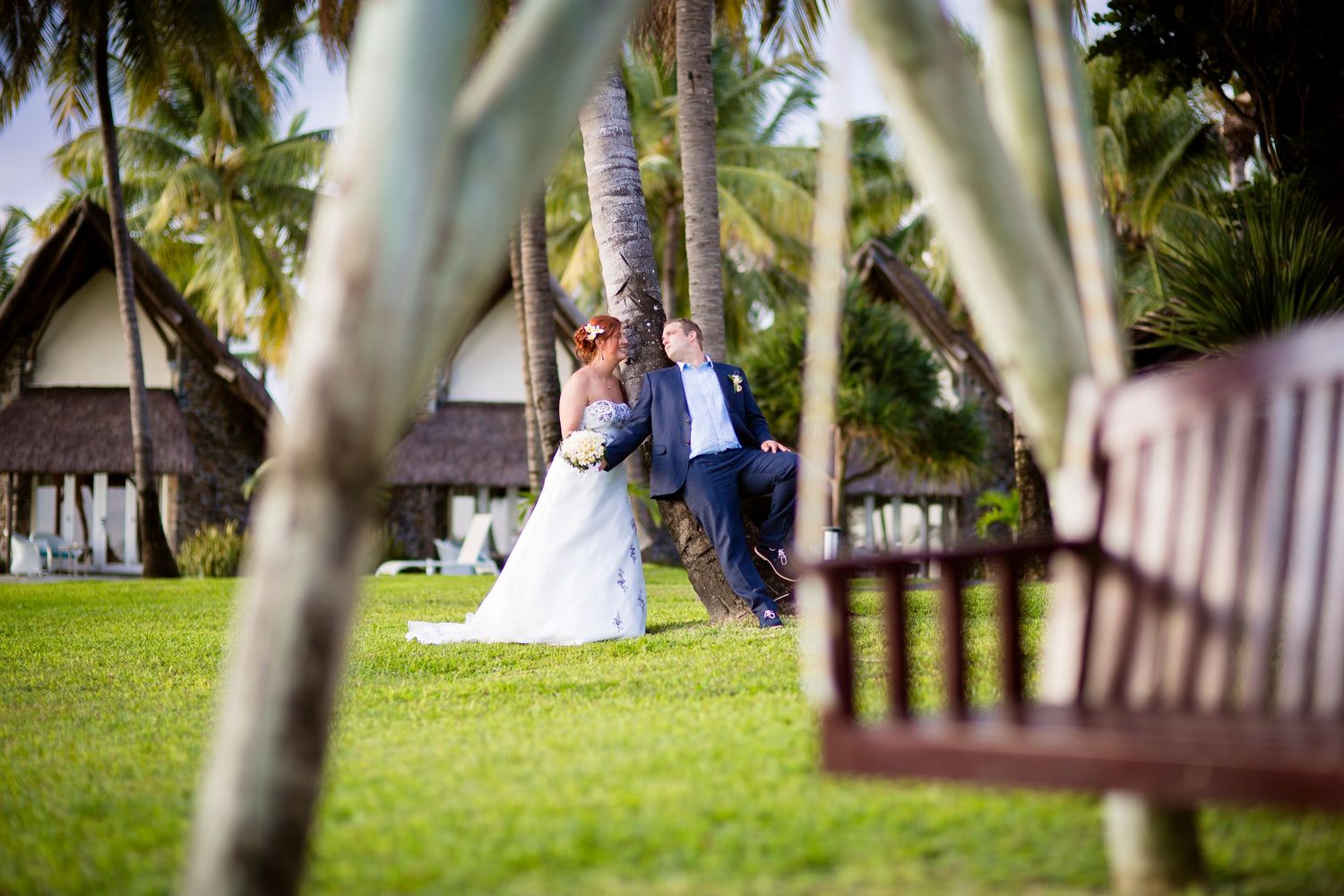 Wedding couple embracing near tropical palm trees at a resort with lush green lawn.
