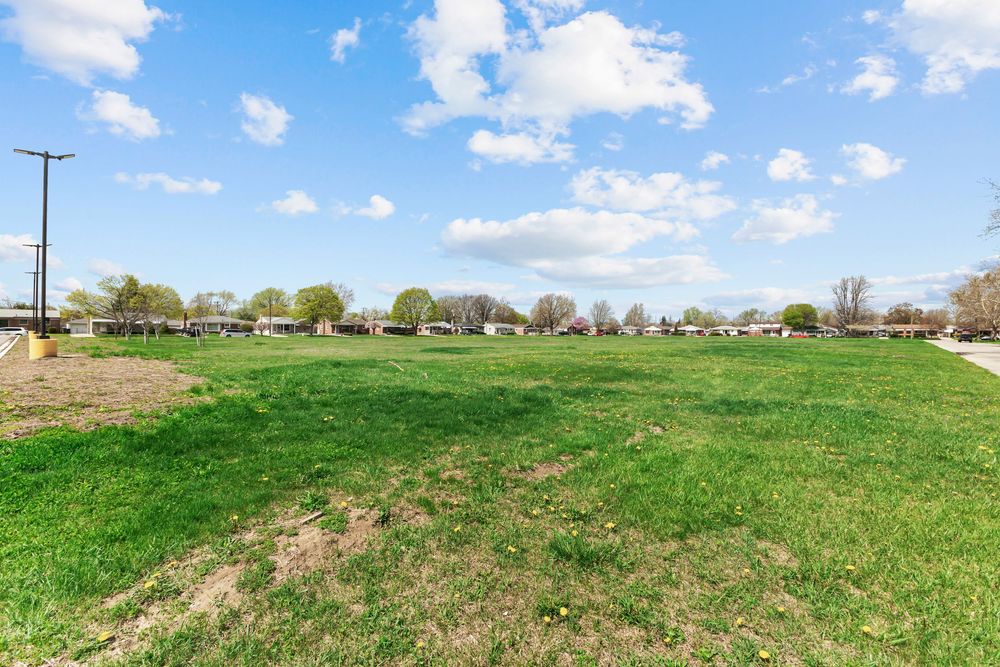 Open green lawn under blue sky with scattered trees and neighboring homes in the background.