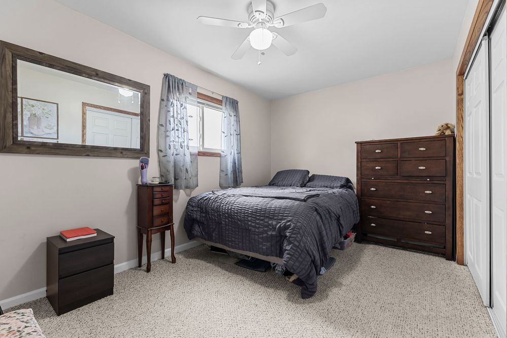 Carpeted bedroom with dark wood dresser, ceiling fan, and natural light from window.