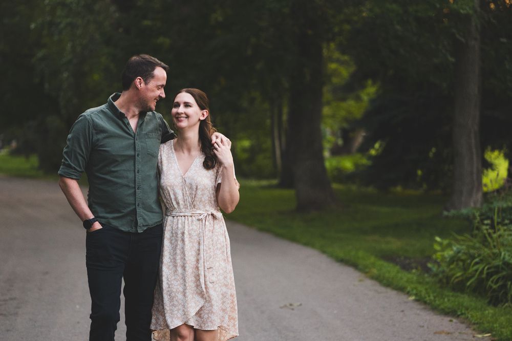 Happy couple walking together on a tree-lined path, woman in lace dress smiling at man in green shirt.