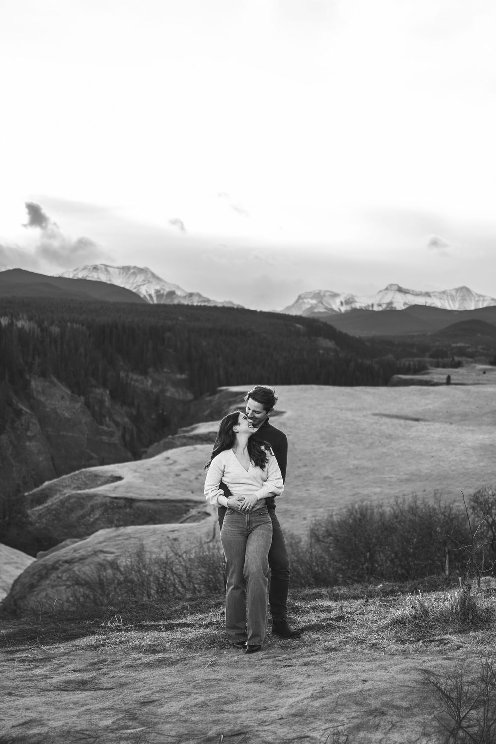 Black and white photo of a couple kissing on a rocky overlook with snow-capped mountains and forest in the background.
