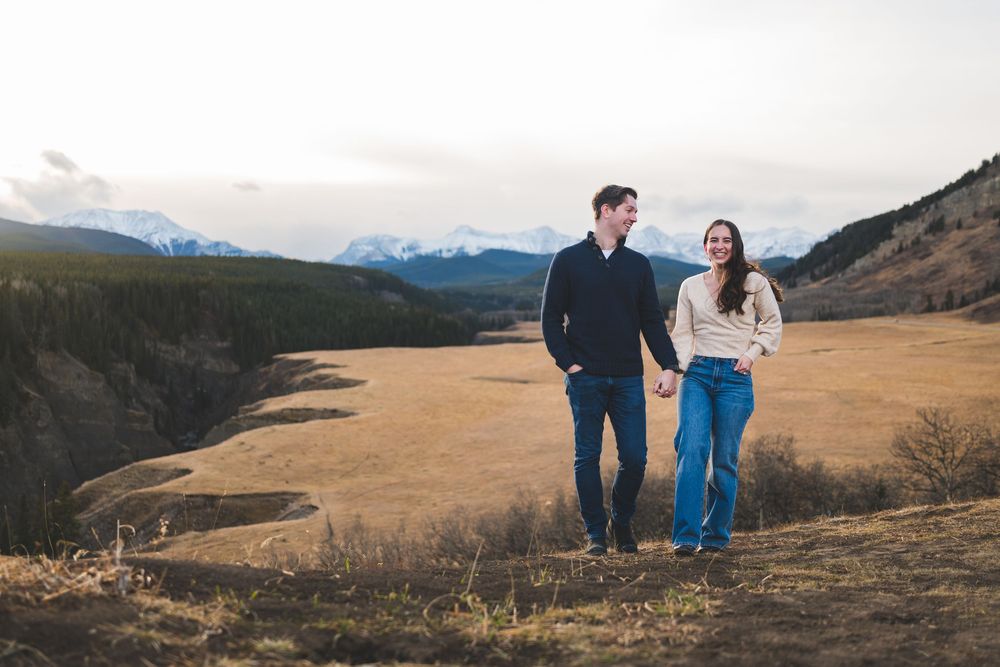 Couple holding hands walking through a golden meadow with snow-capped mountains in the background.