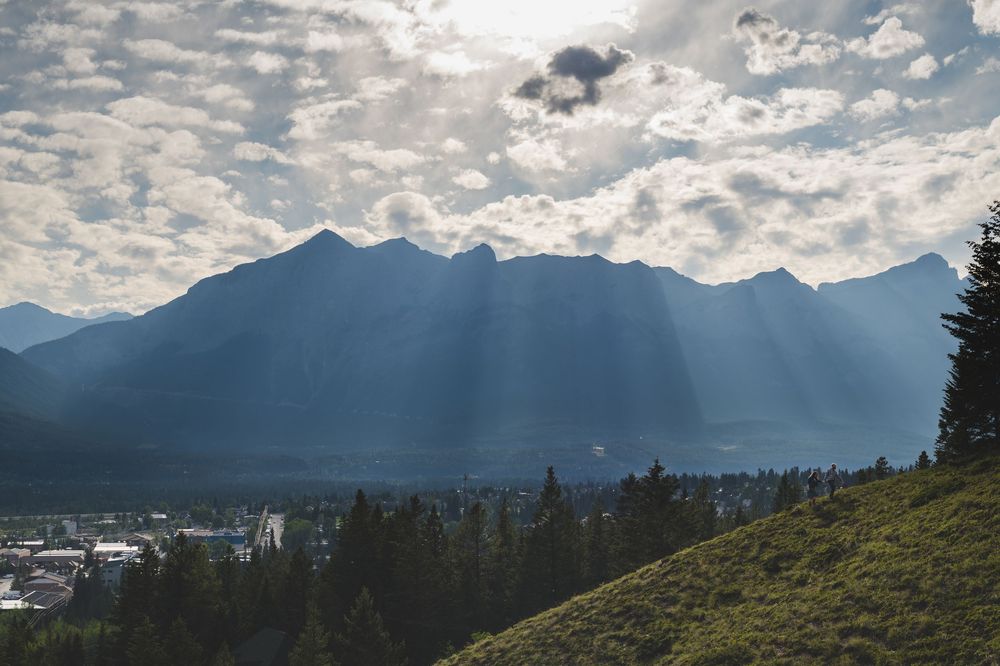 Sunbeams breaking through clouds over a mountain range with a town nestled in the valley below, surrounded by evergreen trees.