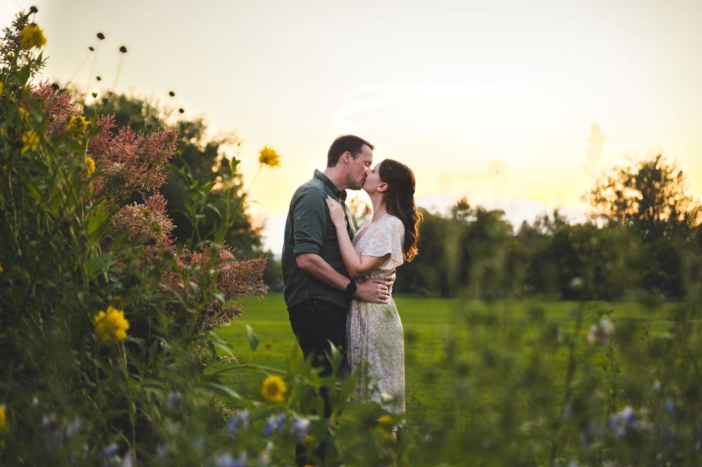 A couple shares a romantic kiss at sunset surrounded by colorful wildflowers in a lush garden field.