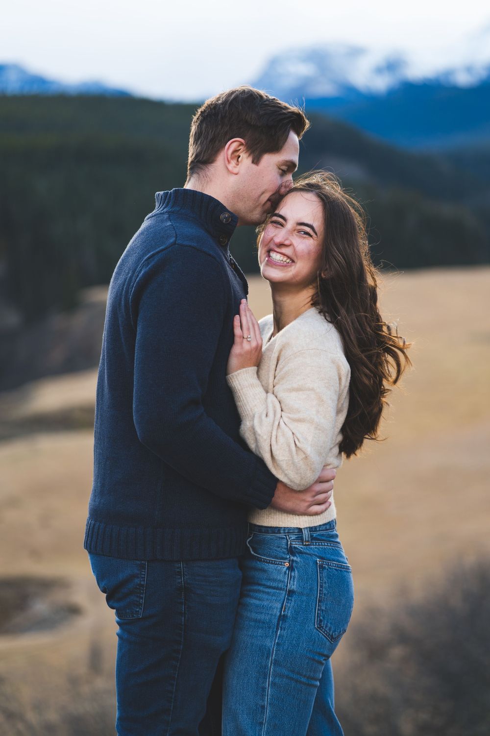 Couple embracing and smiling in a mountain meadow, woman in cream sweater and jeans, man in navy pullover.