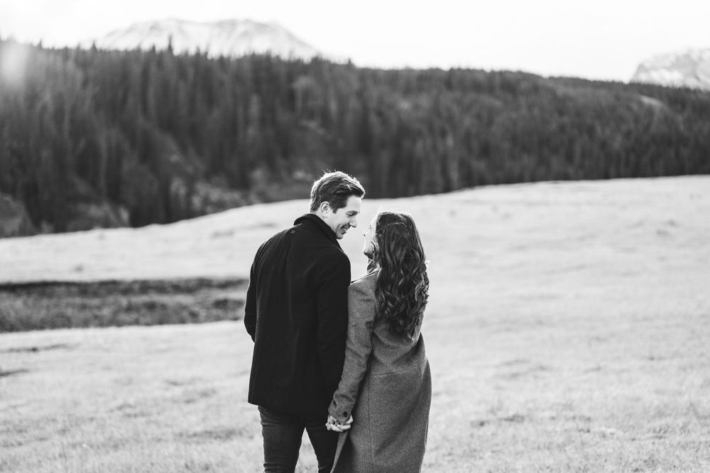 Black and white photo of a couple holding hands in a mountain meadow, sharing a romantic moment with forested hills behind.