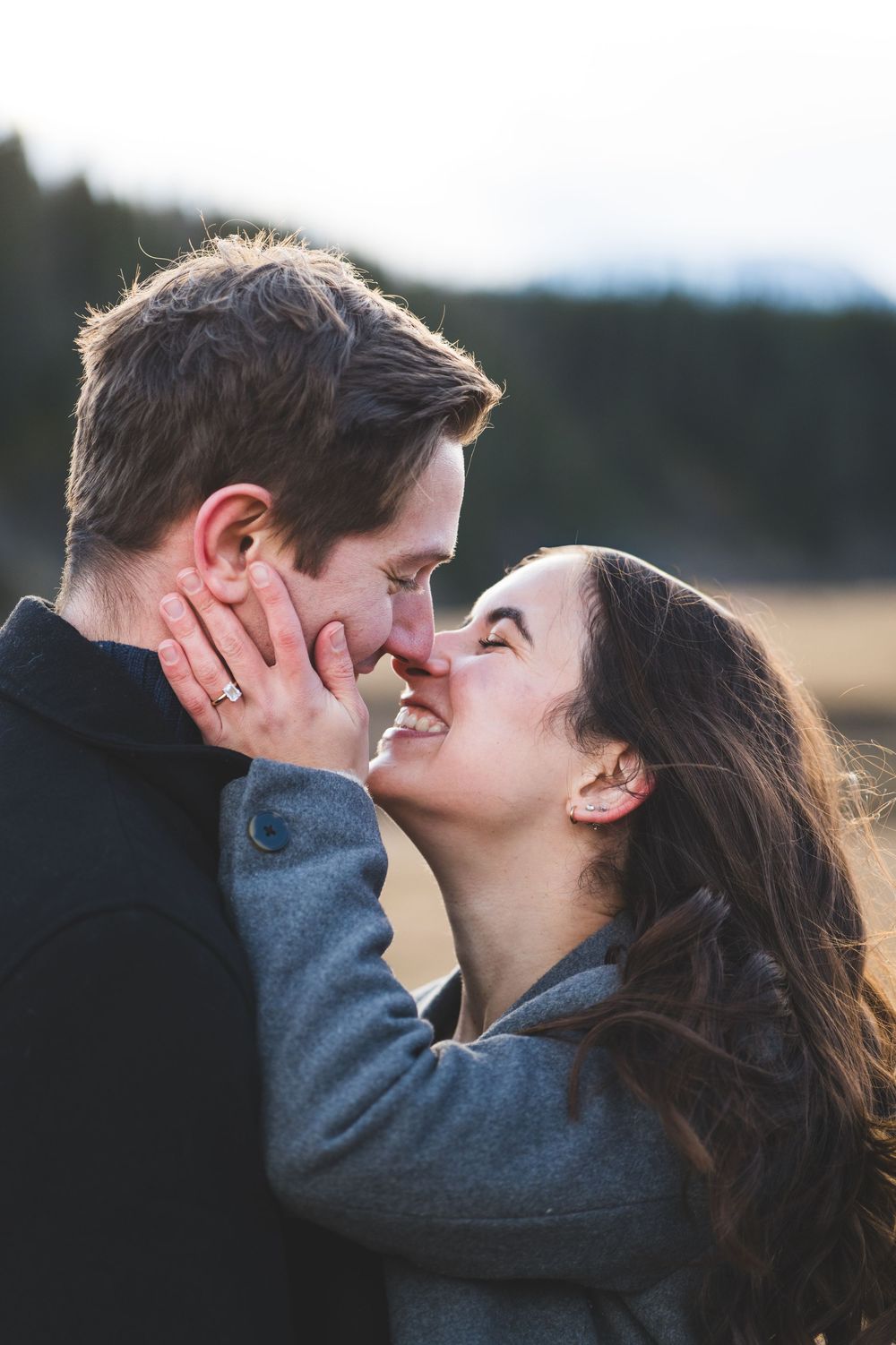 Happy couple about to kiss outdoors, woman holding mans face, both smiling, mountains in background.