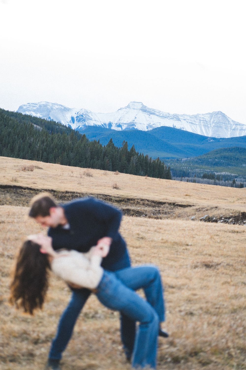 Romantic couple sharing a dip kiss in a golden meadow with snow-capped Rocky Mountains in the background.
